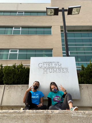 Two masked students giving V-sign in front of Guelph-Humber building sign