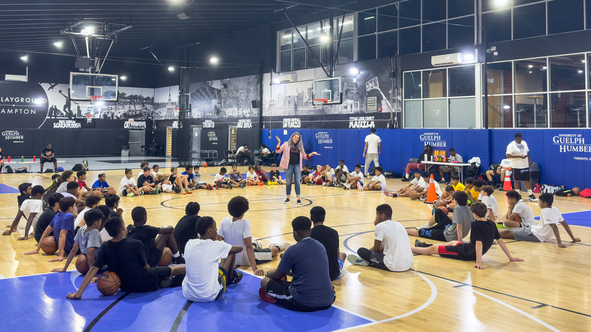Nikki Martyn speaks to kids sitting around in a circle in the middle of an indoor basketball court