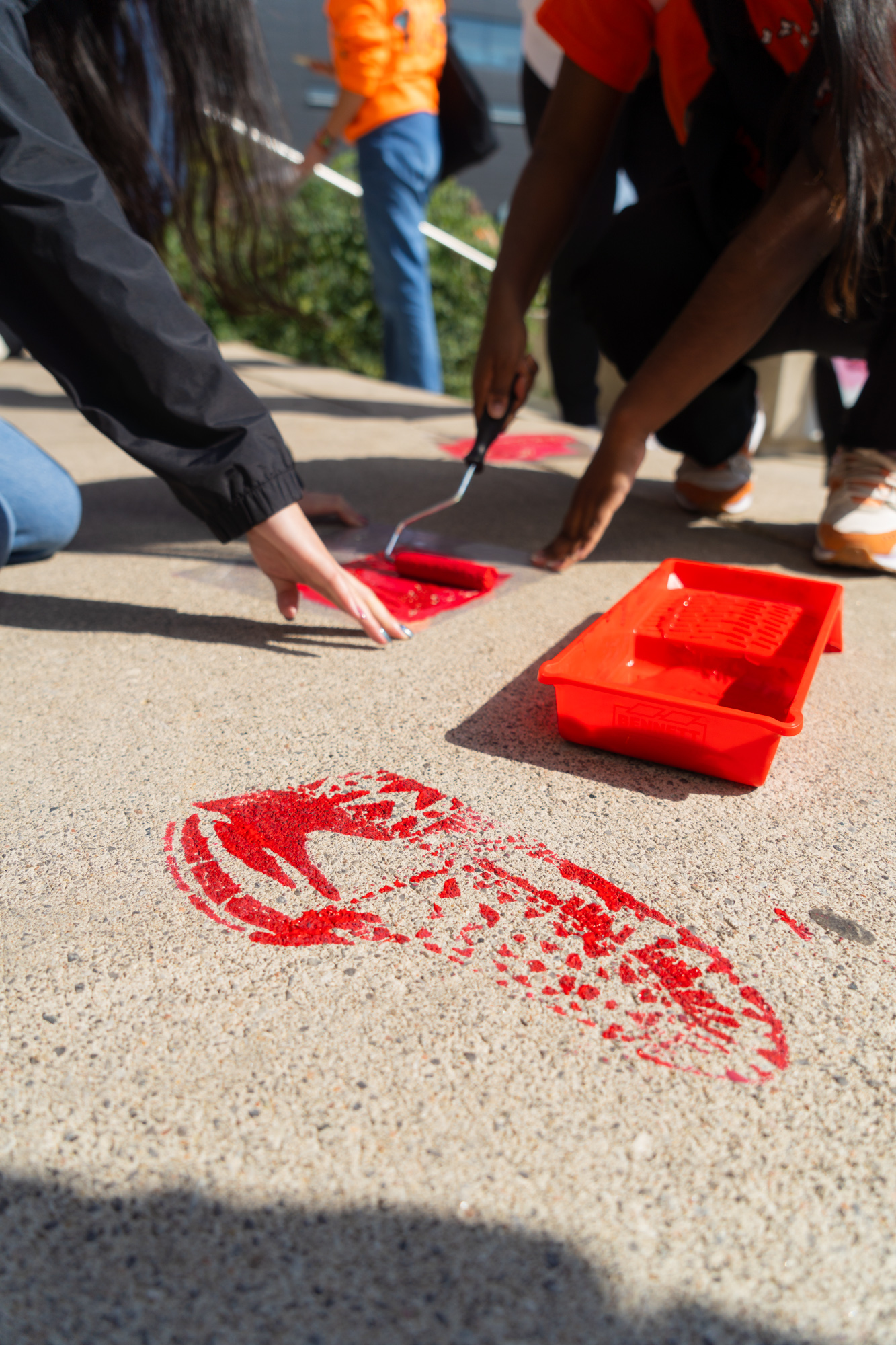 A red paint-stencilled moccasin with two students in the background stencilling another moccasin.