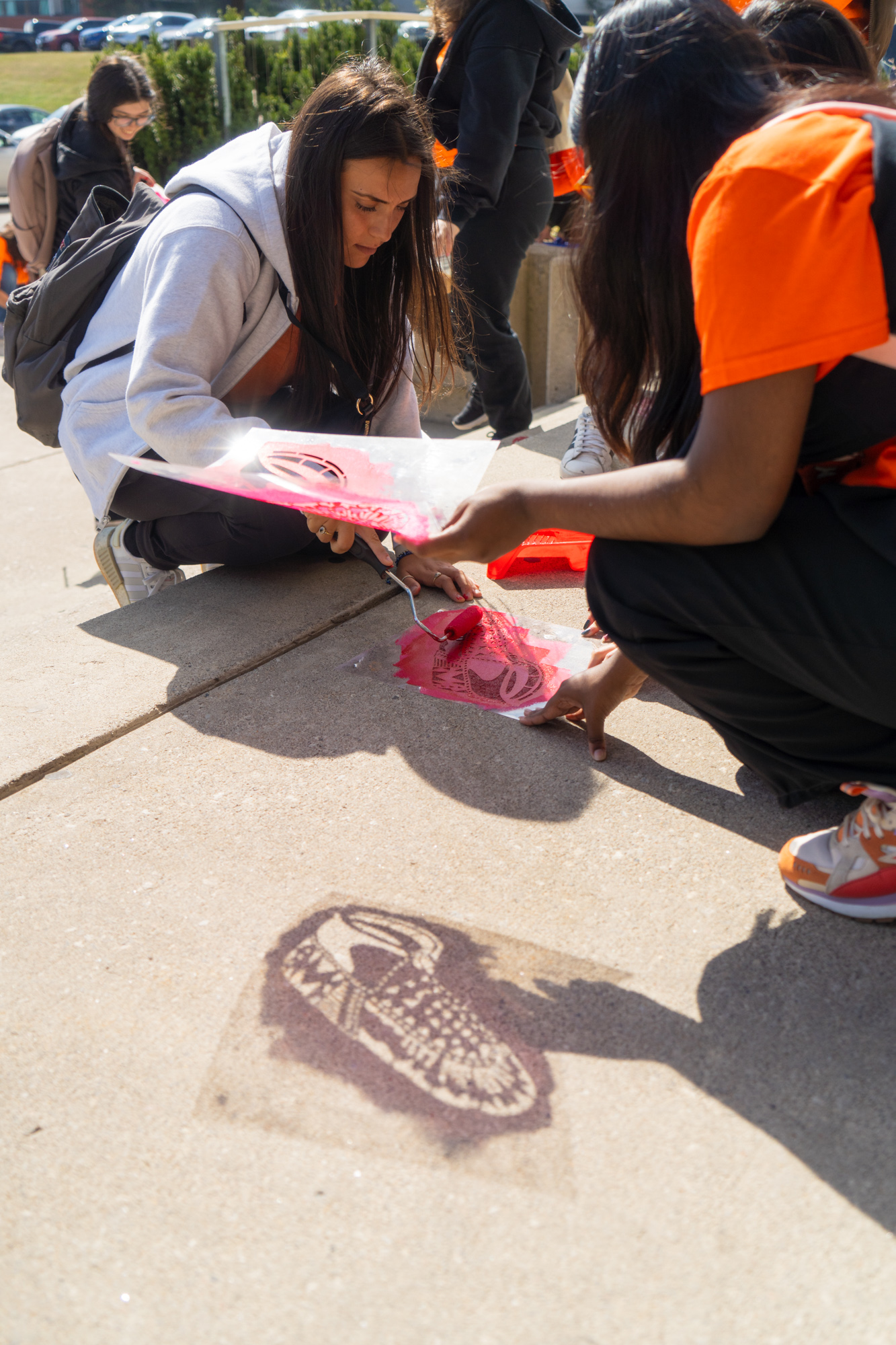 A student holding up a moccasin stencil casts a shadow in the foreground while a another rolls paint onto another stencil