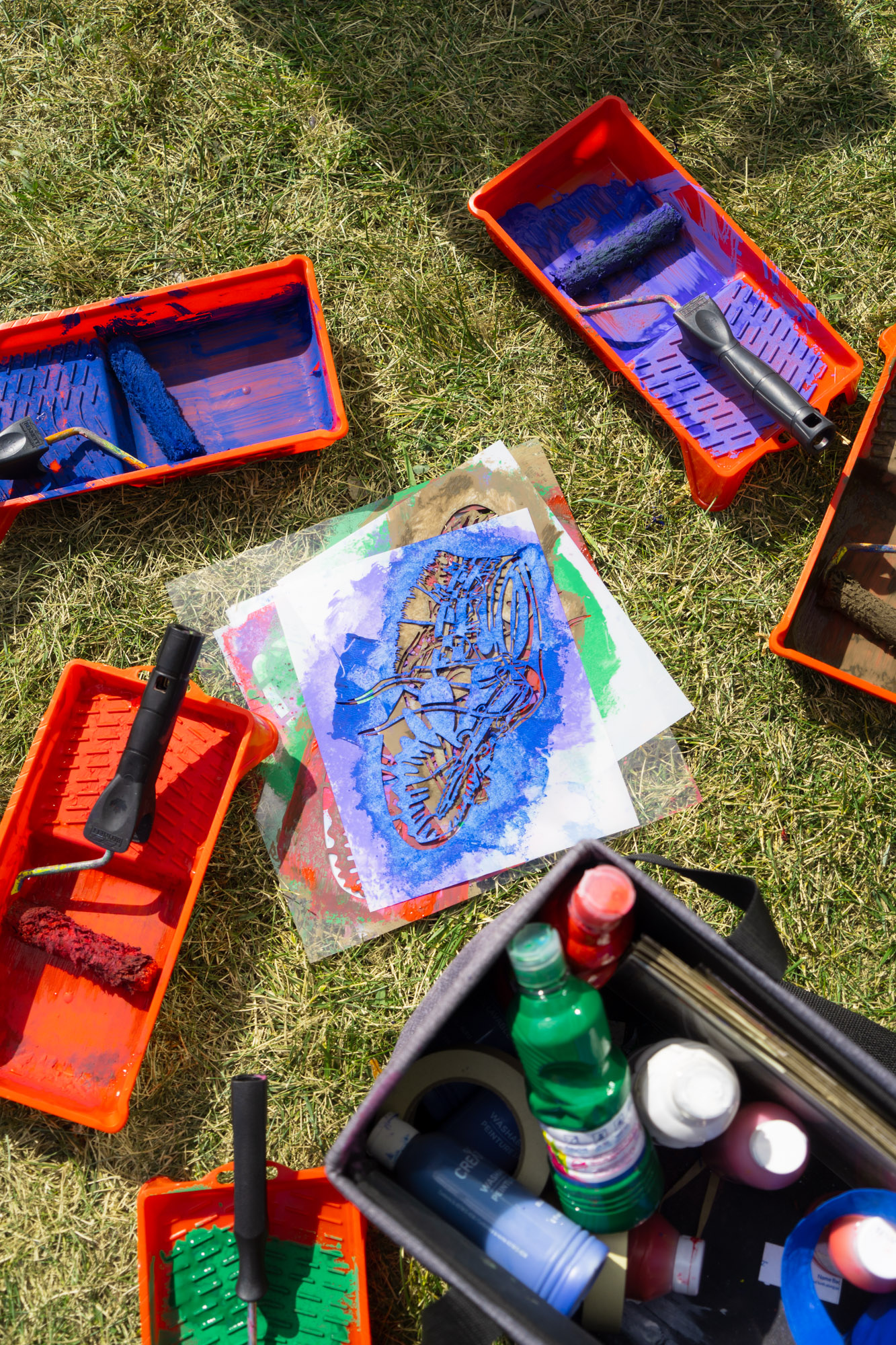 A pile of coloured moccasin stencils surrounded by paint trays, paint rollers, and a box of paint tubes