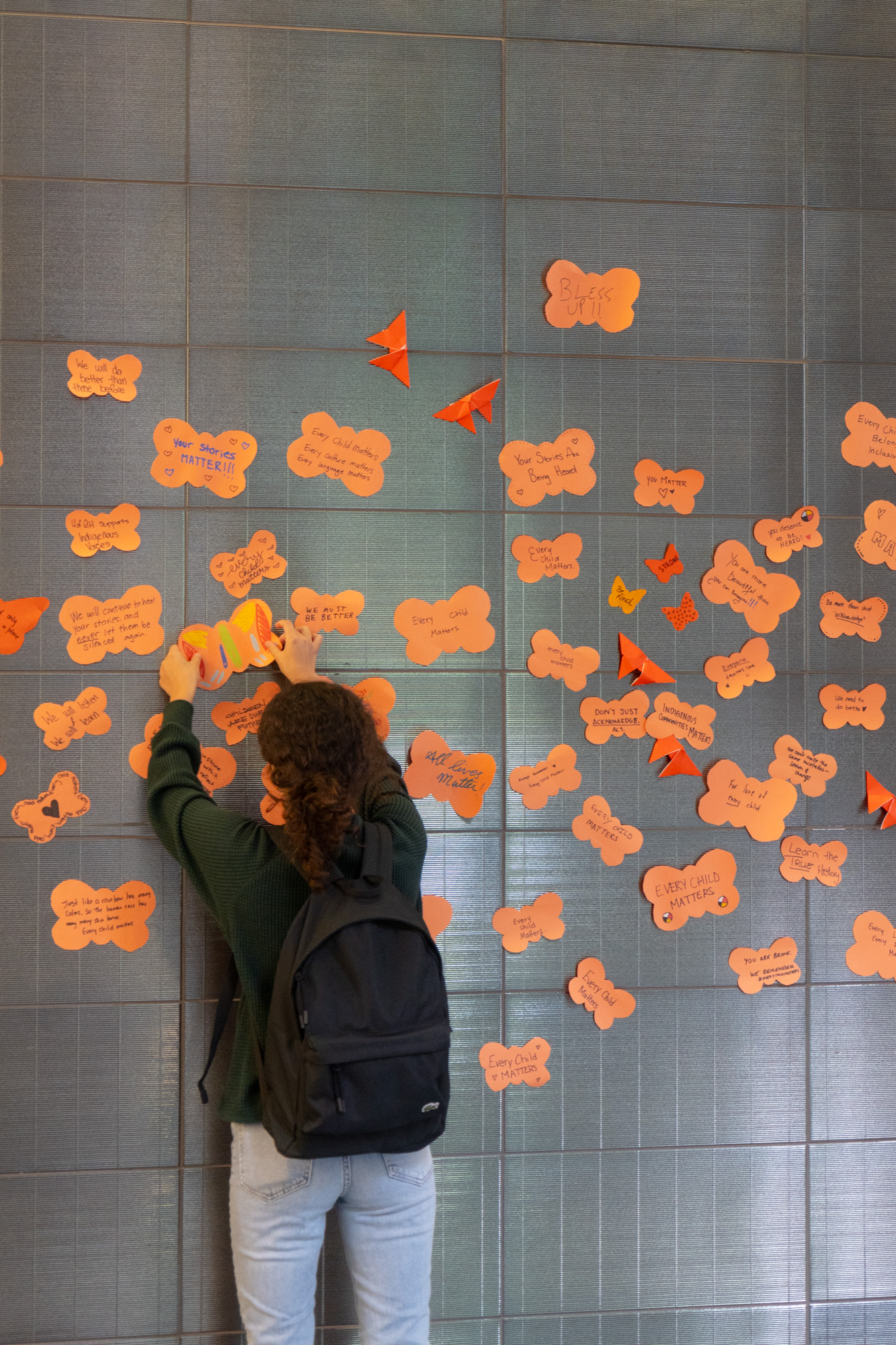 A person places an orange butterfly-shaped paper on a wall amongst other similarly shaped paper and origami