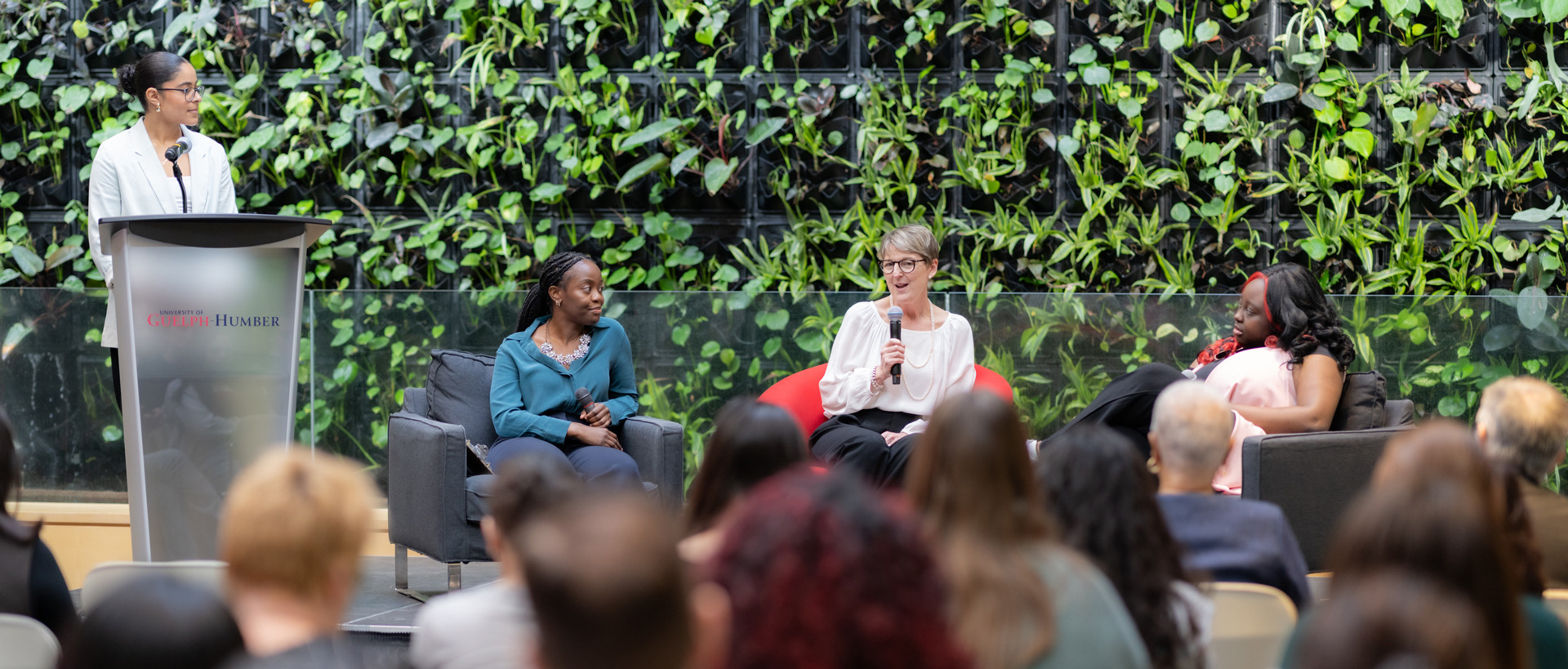 Three individuals sitting on couches as panelists while one is standing at a podium