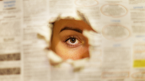 An eye peering through a newspaper