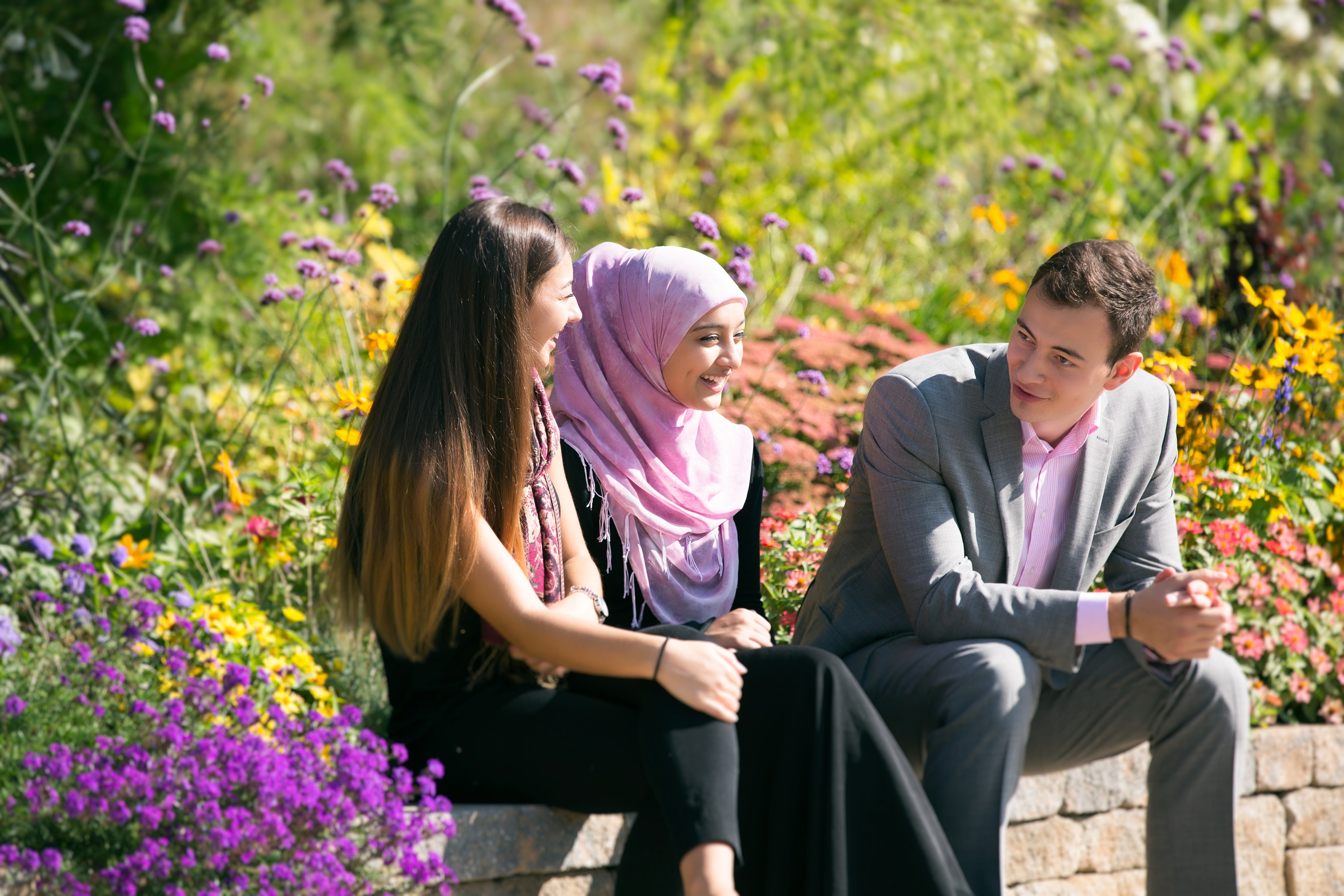 Three students sitting on a bench in nature with plants and flowers behind them
