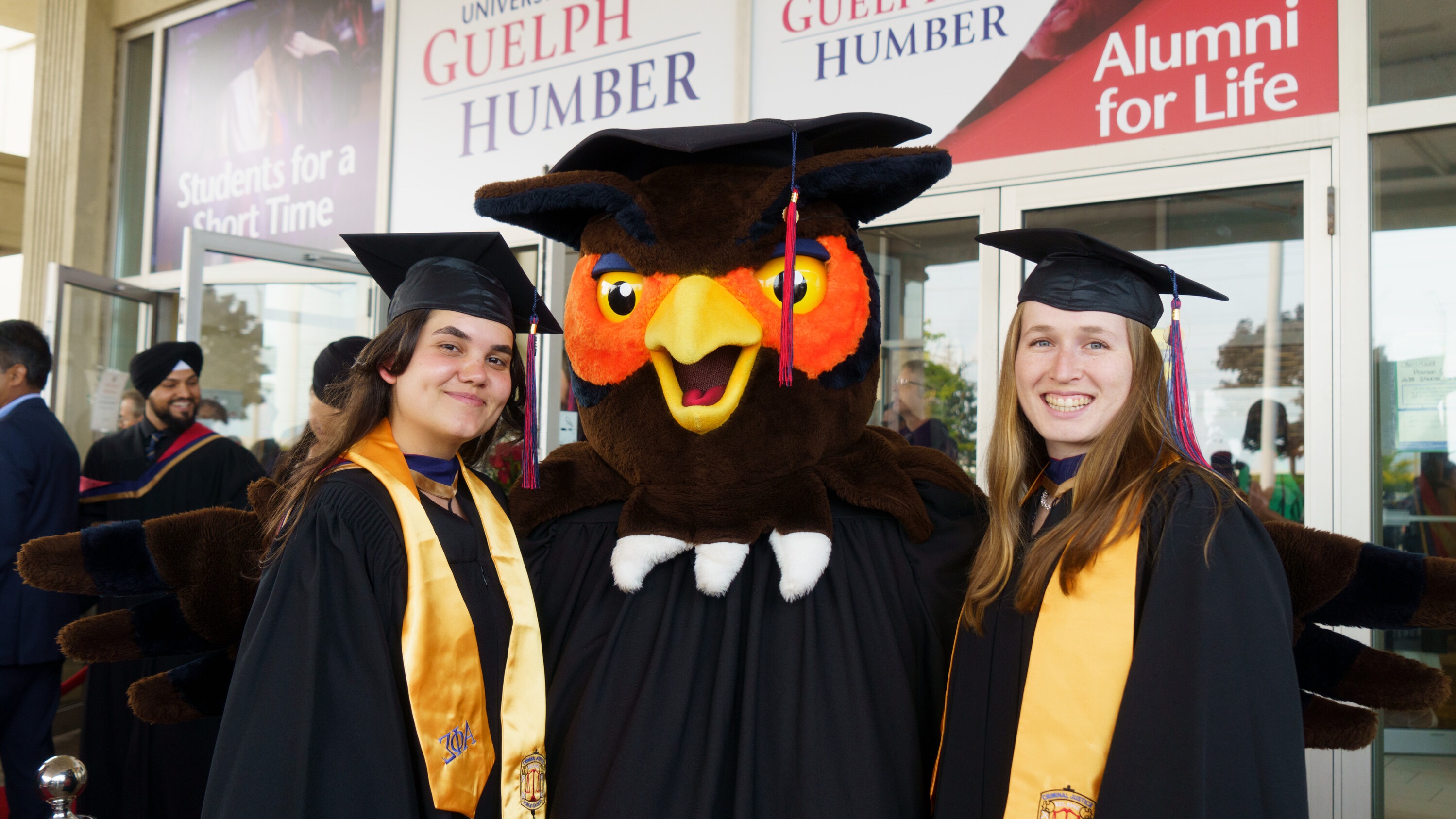 Two students in convocation regalia with Swoop the University of Guelph-Humber owl mascot, also in regalia
