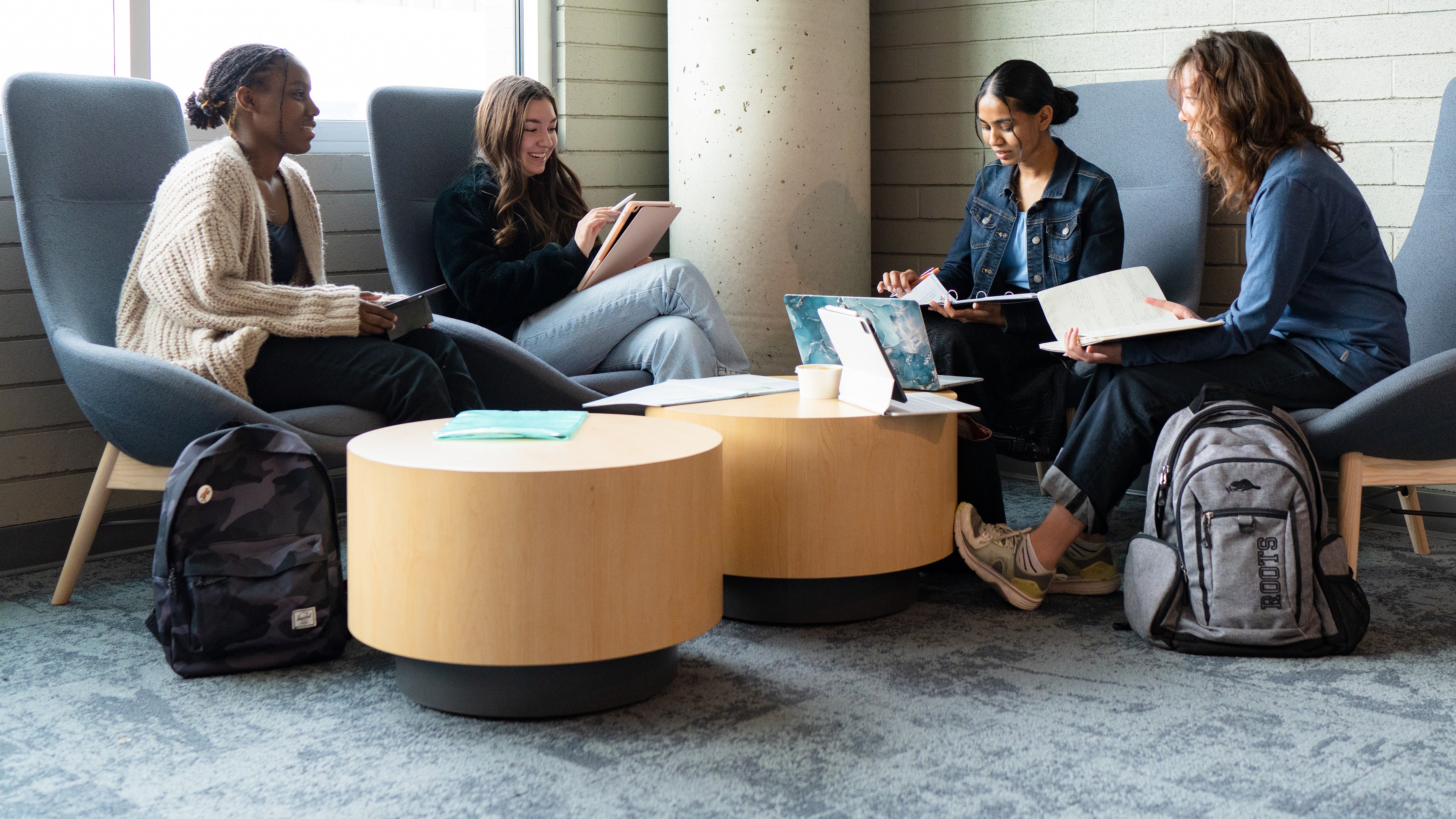 Group of four students sitting in comfy chairs with round coffee tables doing work