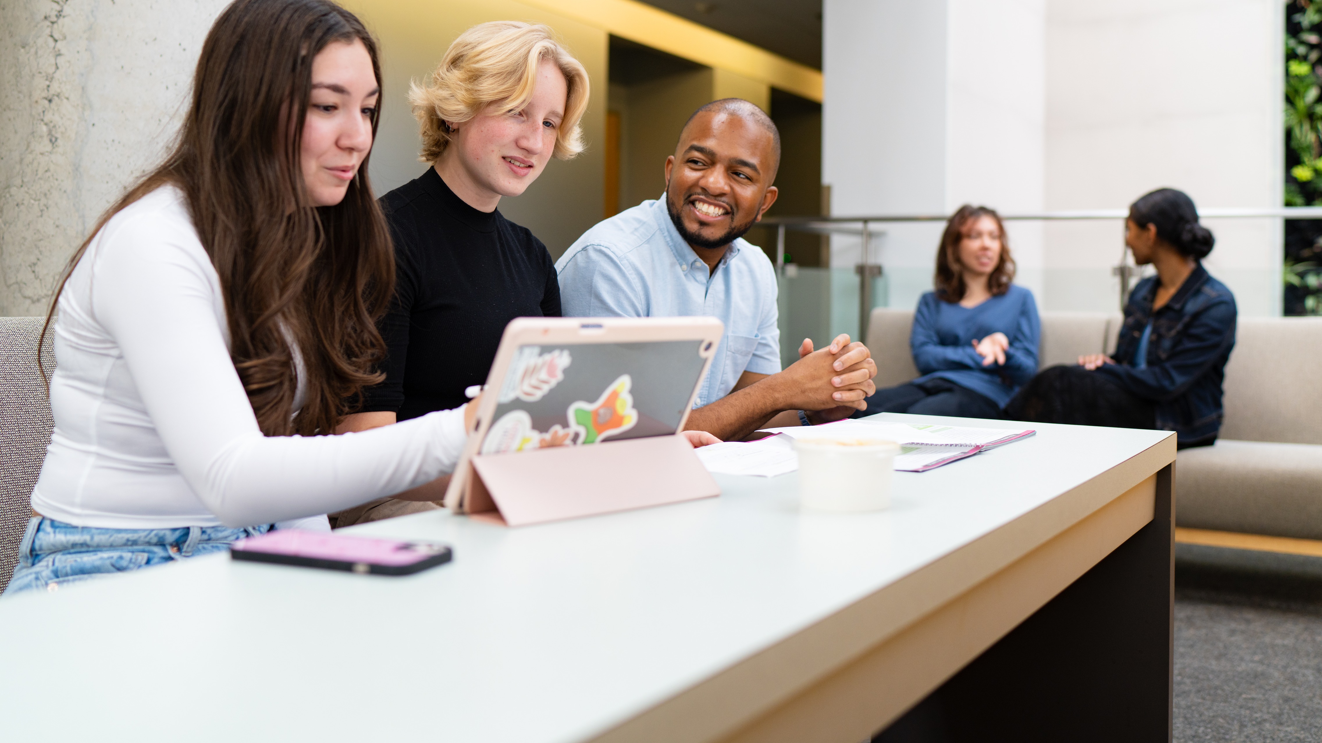 Three students sitting at desk looking at laptop with two students in the background