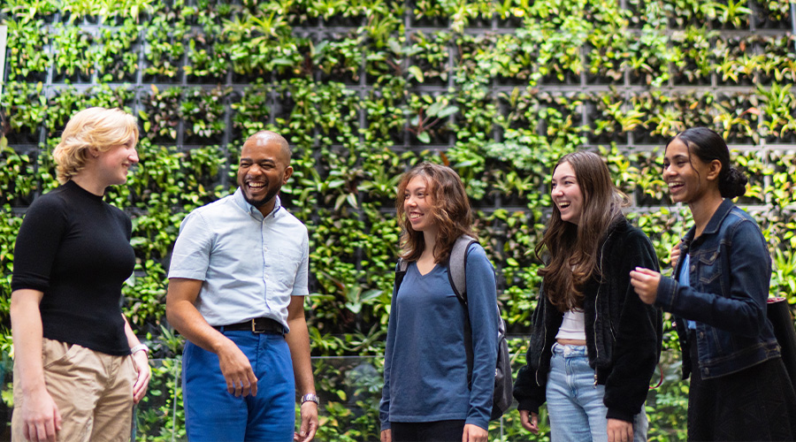Five people laughing in front of a plant wall