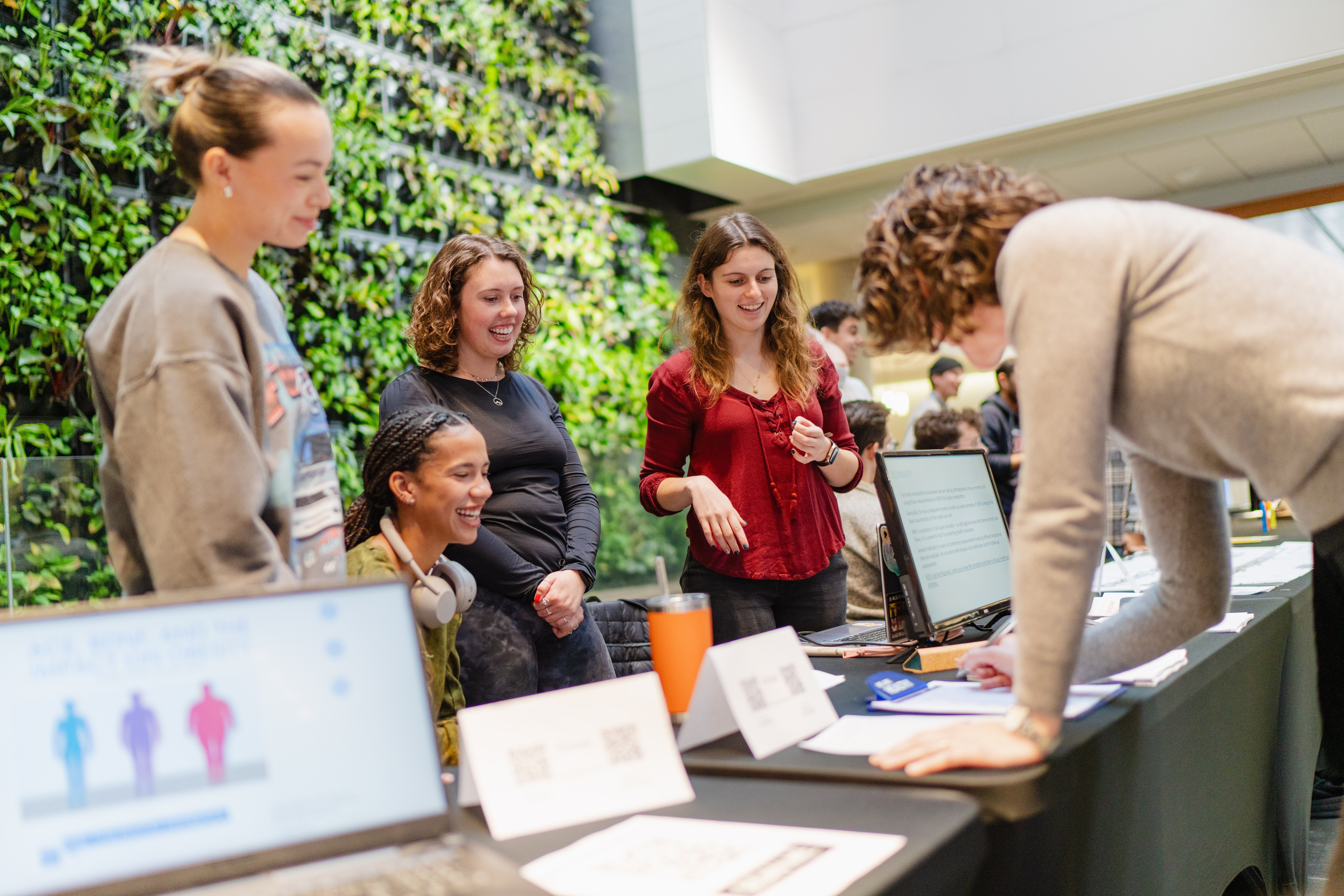 Students presenting to a professor at a table in front of the plant wall