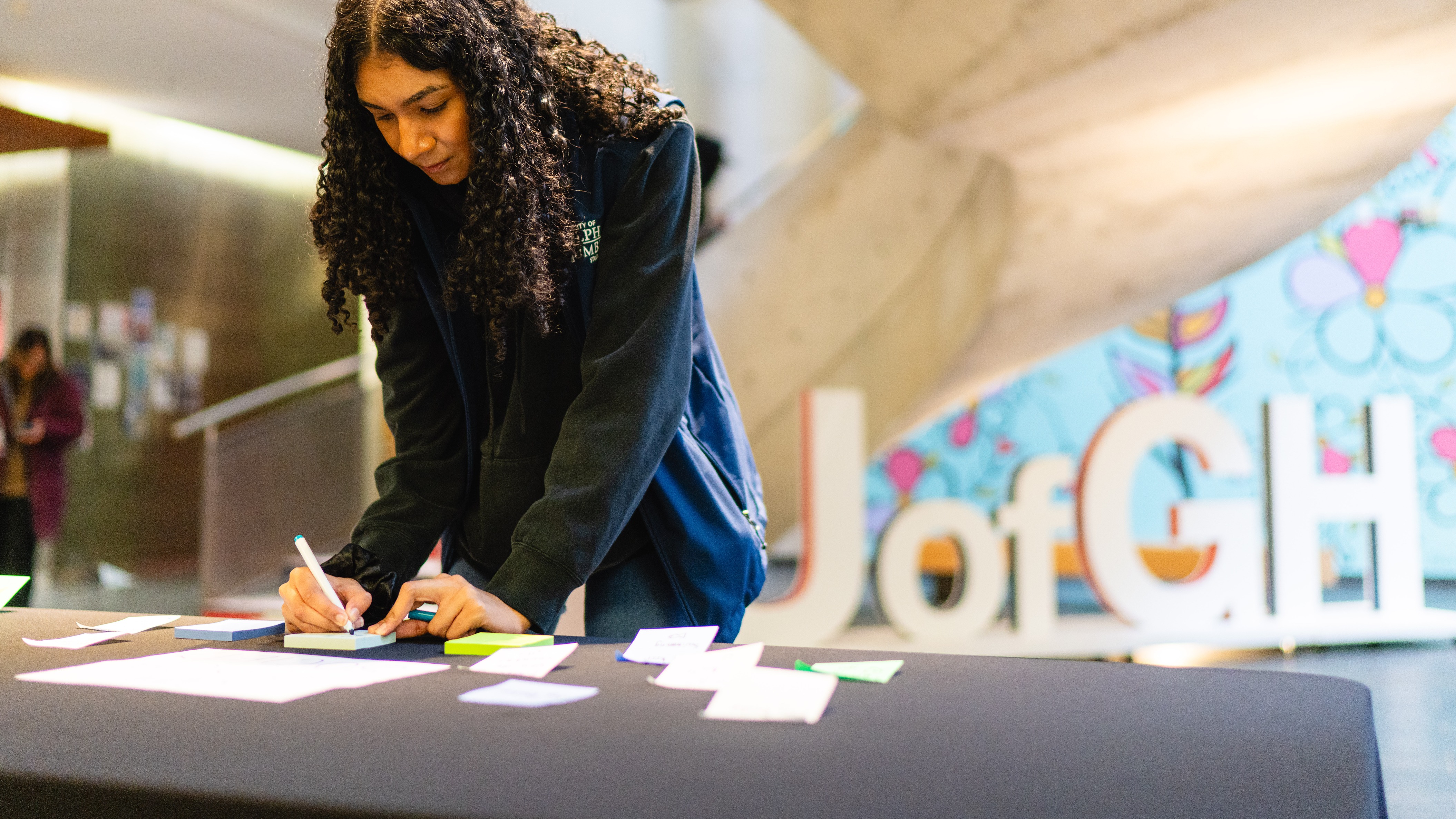 student standing at desk writing on a card with large U of GH letters behind