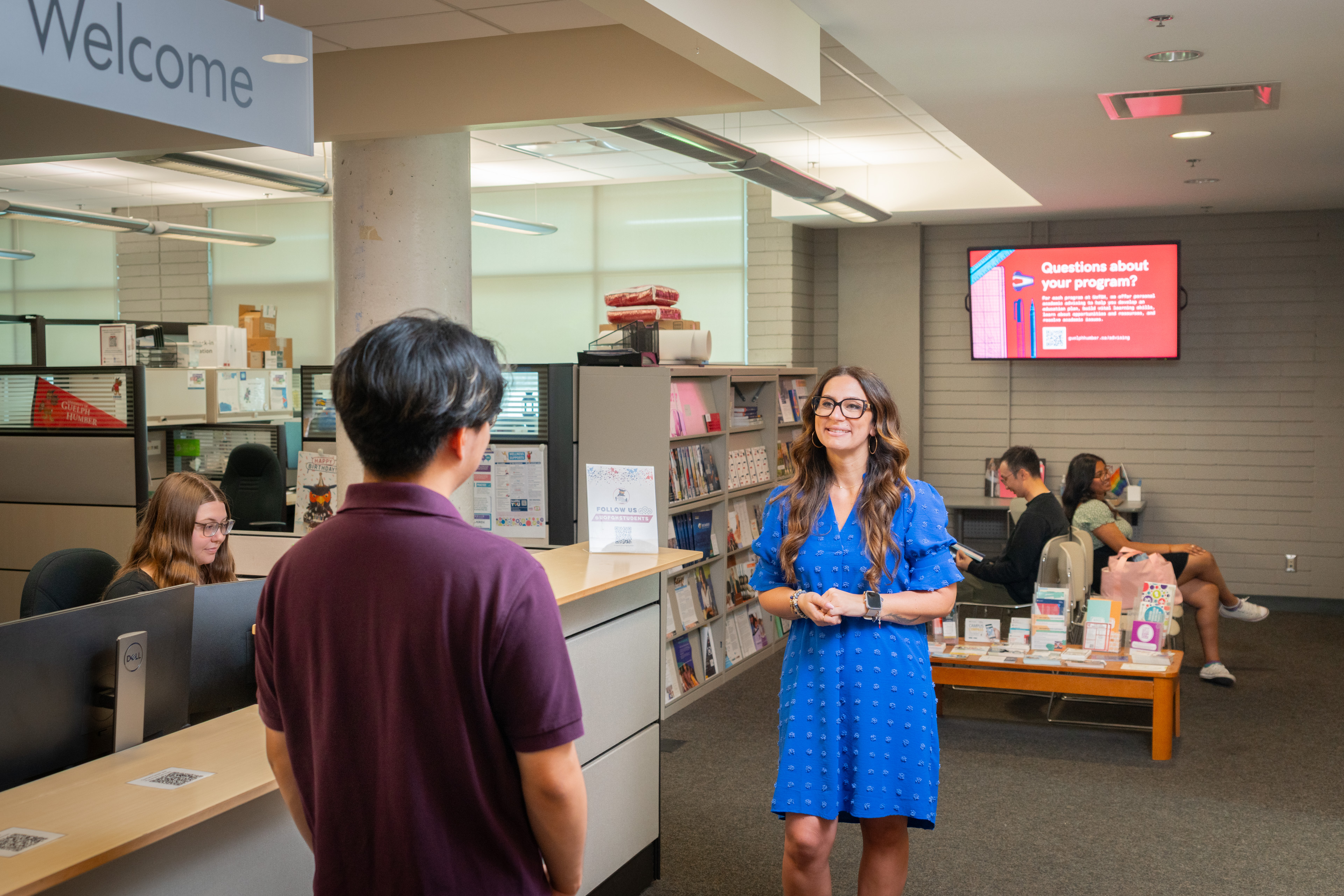 Two people talking in reception area with person at desk and two people in seats in the background