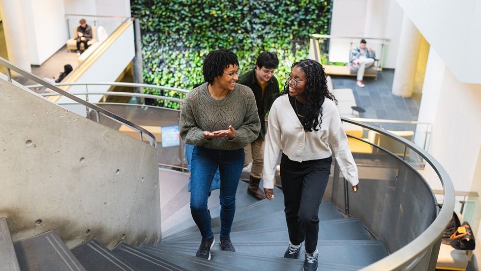 Three students walking up the spiral staircase with the plant wall in the background
