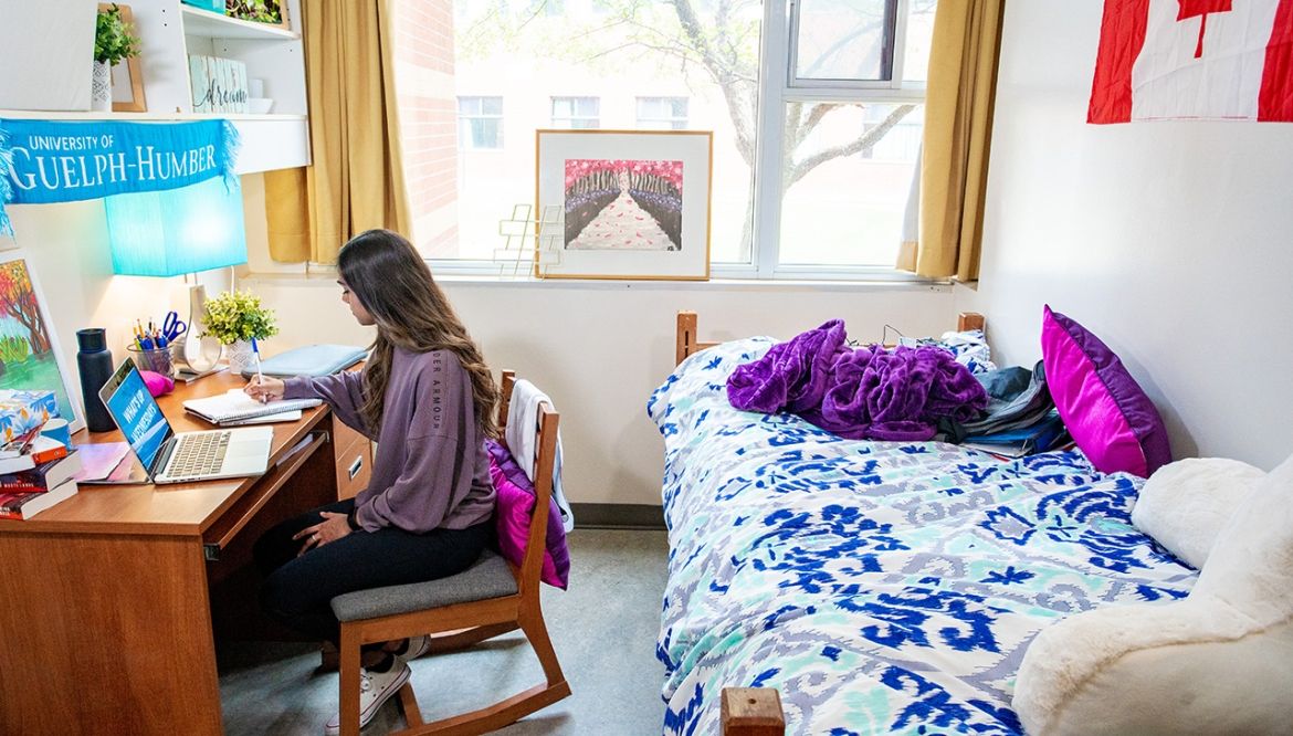 A female student sits at a desk and works on her laptop in a bright dorm room.