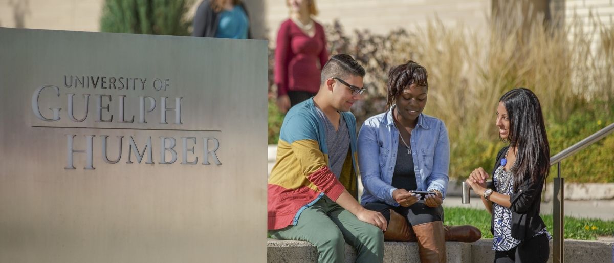 Three students sitting on ledge outside of the GH building next to the sign that says University of Guelph-Humber