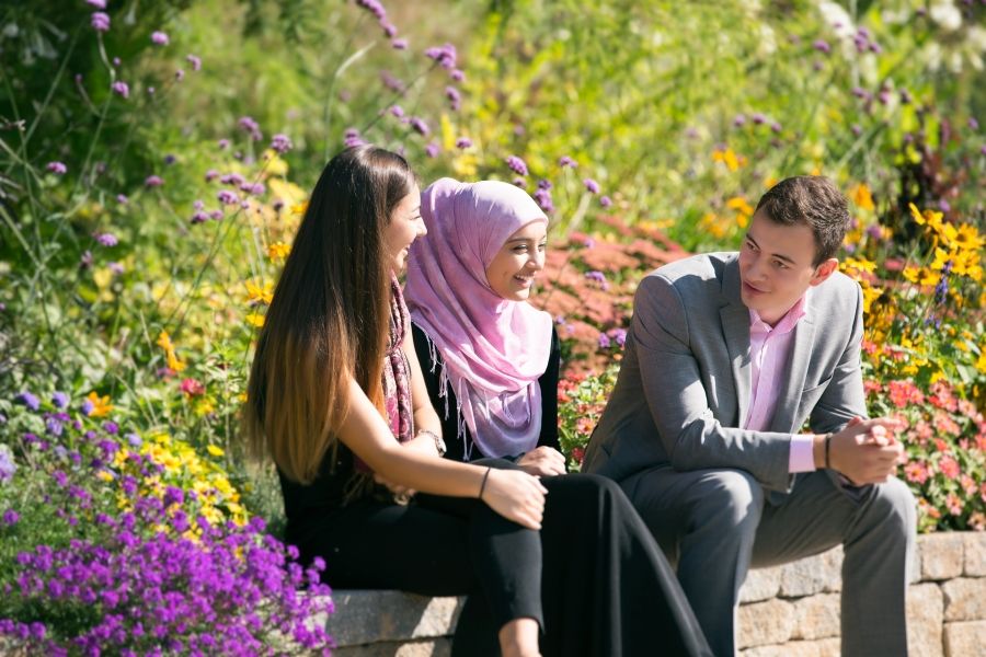 Three students sitting on a bench in nature with plants and flowers behind them