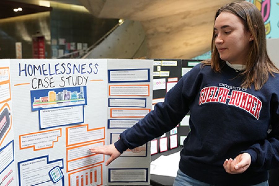 Female student with U of GH sweater in front of poster