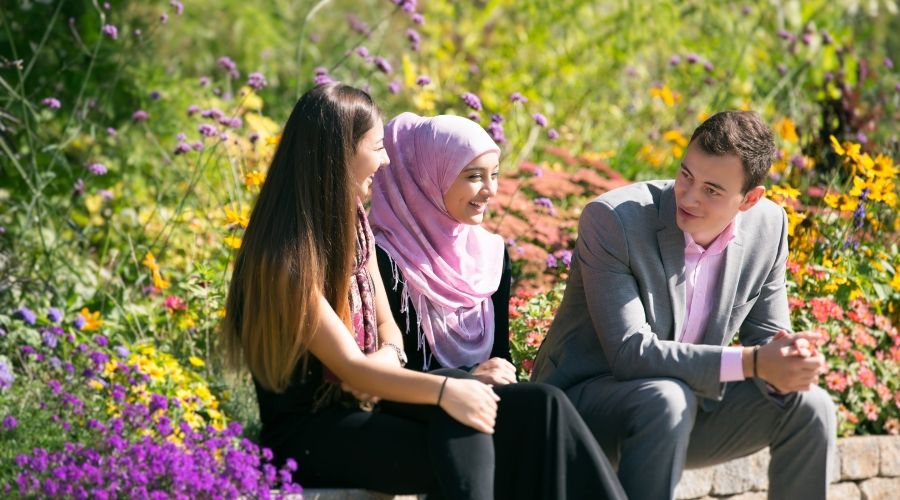 Three students sitting on a bench in nature with plants and flowers behind them