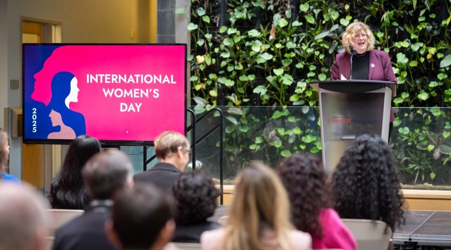 Dr. Anne Marie Vaughan speaking in front of an audience on a stage in front of a plant wall and a TV displaying &quot;2025 International Women's Day&quot; on the screen.