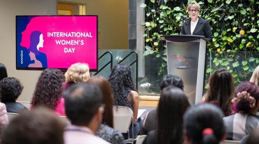 Dr. Melanie Spence-Ariemma speaking at a podium on a stage in front of a plant wall with a TV displaying, &quot;2025 International Women's Day&quot; to the right of the stage.