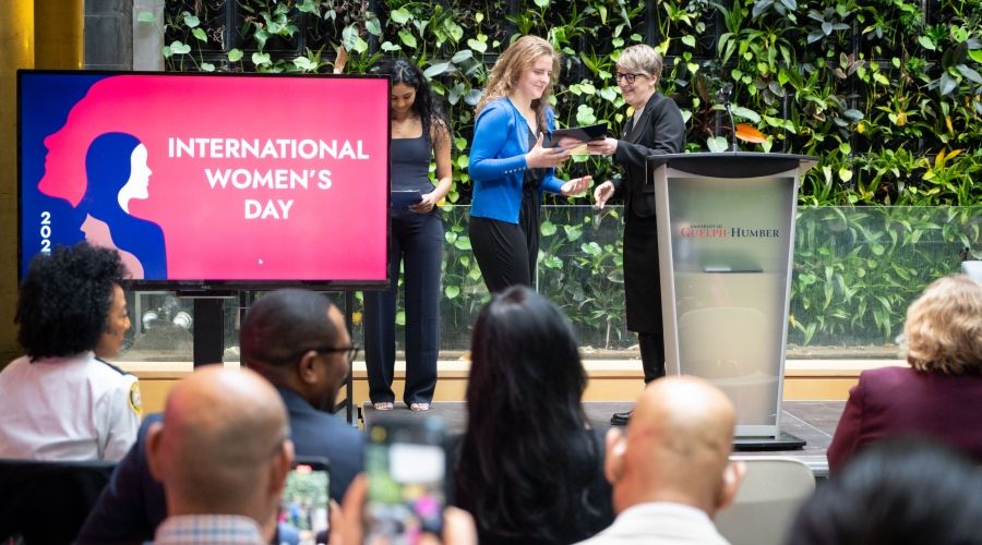 Someone receiving an award from another person while a TV screen on their right displays, &quot;2025 International Women's Day&quot;