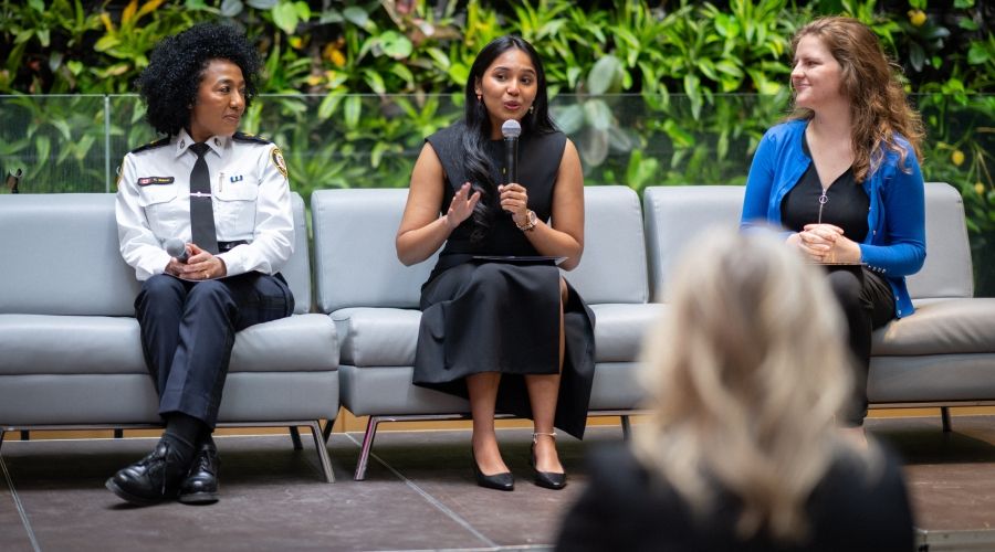 Someone speaking into a microphone while two others look at them, one person on either side. They are sitting on grey couches with a plant wall behind.