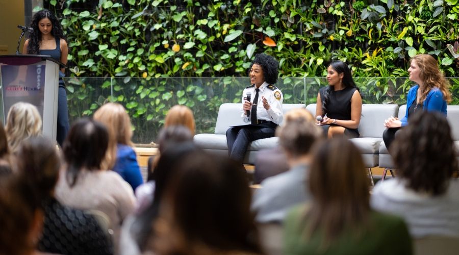 Someone speaking into a microphone while two others look at them. They are sitting on grey couches with a plant wall behind. There is also one person standing a podium off the left side.