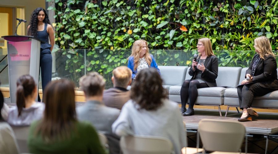 Someone speaking into a microphone while two others look at them, one person on either side. They are sitting on grey couches with a plant wall behind, and an audience in front of them.