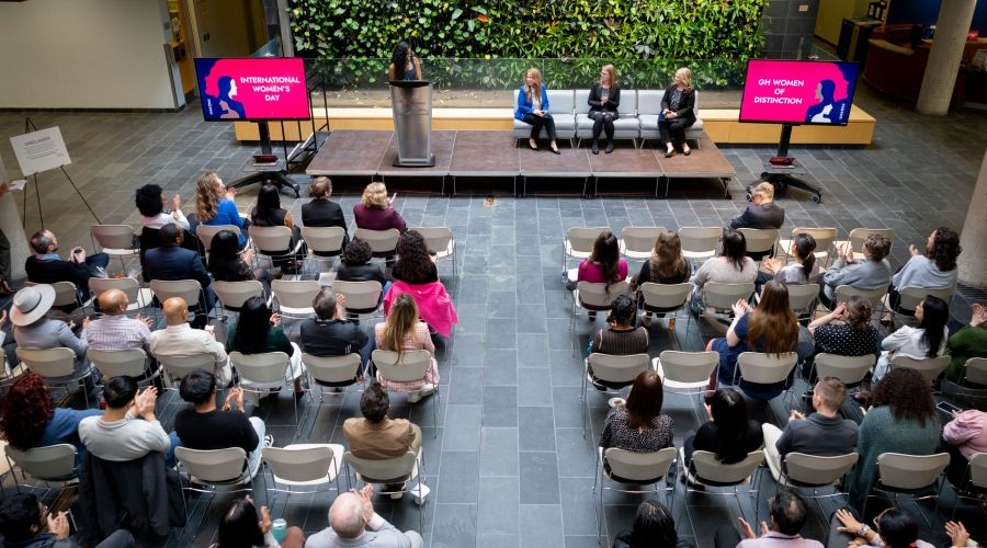 An aerial view of the Guelph-Humber atrium filled with an audience sitting in chairs while they watch three panelists sitting in grey couches and one moderator at a podium speak.
